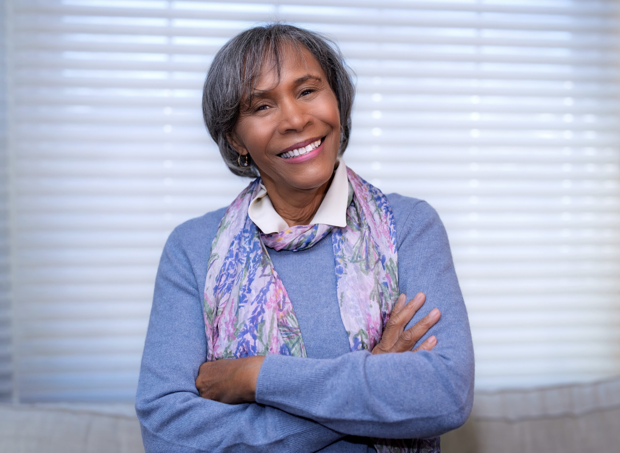Smiling older woman with short gray hair, wearing a blue sweater and floral scarf, stands with arms crossed in front of a window with blinds.